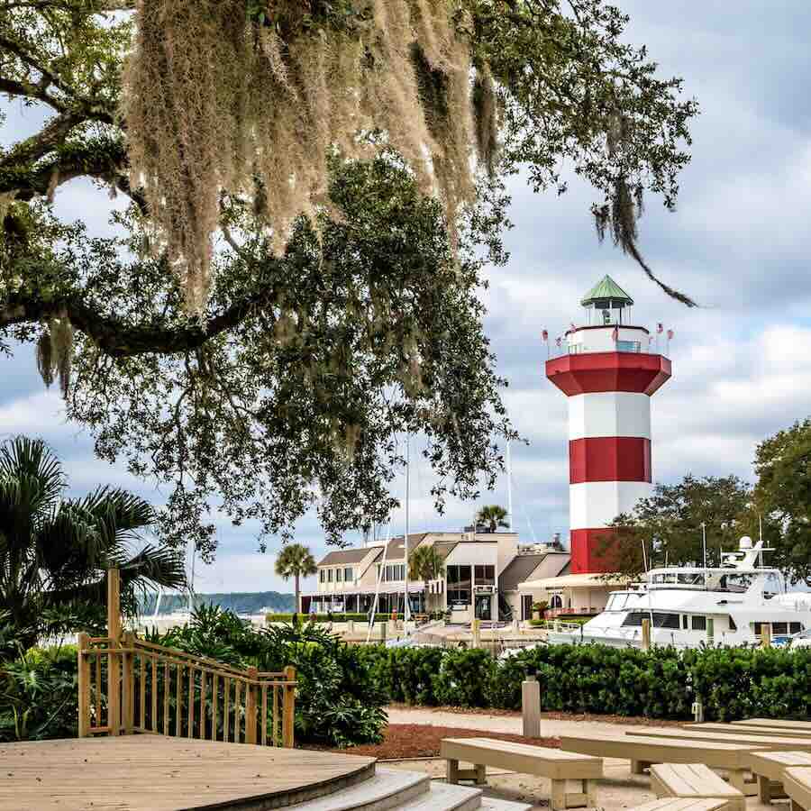 Lighthouse and boats at marina with trees nearby.