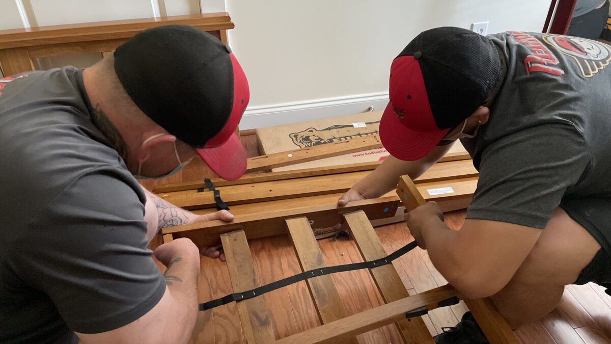 Two men assembling wooden bed frame indoors.