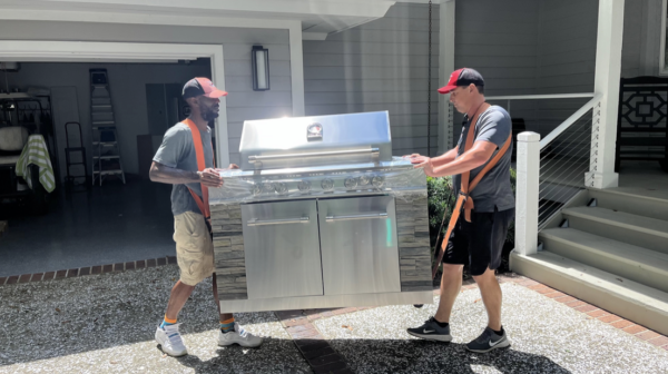 Two people moving a stainless steel outdoor grill.